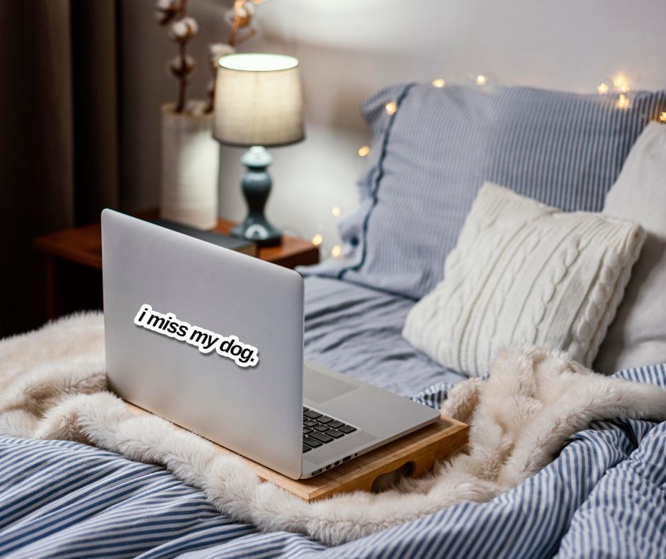 Laptop on a wooden stand with 'I miss my dog' sticker on a bed with pillows and a lamp in the background.